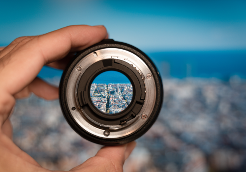 A large city in the distance, being viewed through a small camera lens being held up by a person’s hand offscreen.